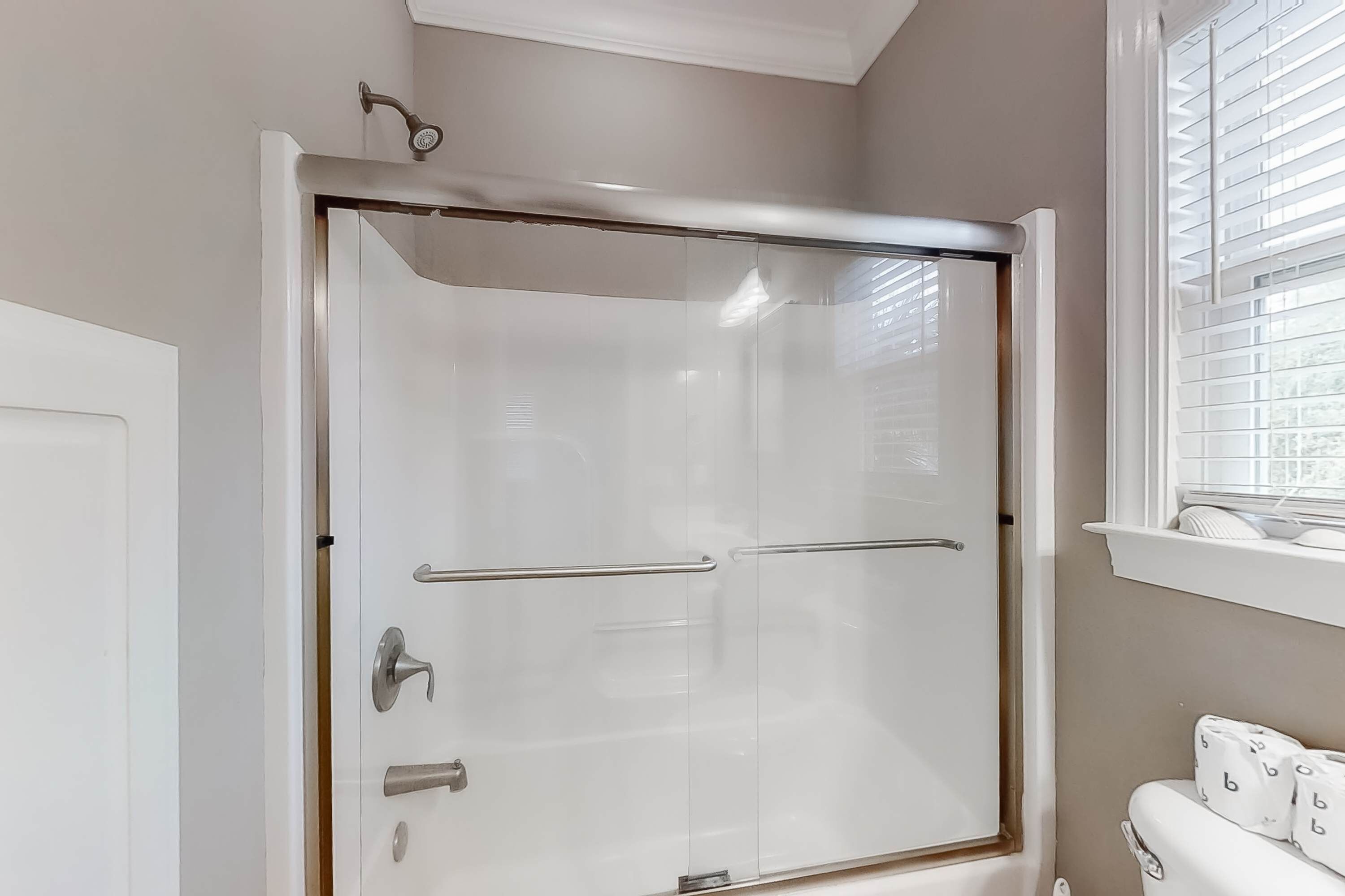 Ducha con puerta de cristal, toallero y ventana con persianas.