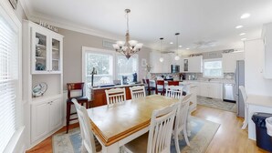A modern kitchen with white cabinetry, a dining table with chairs, a chandelier, and a view of the outside through large windows. - Fantastic House 2 Blocks From Beach w/Private Pool, Free WiFi, Central AC, & W/D (Edisto Beach)
