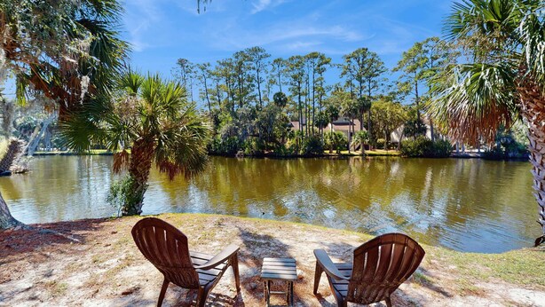 A serene outdoor seating area with two chairs and a small table, overlooking a calm lake with trees and a distant building. - Beautiful home with natural light, lagoon views - Community Hot tub & Pool (Edisto Beach)