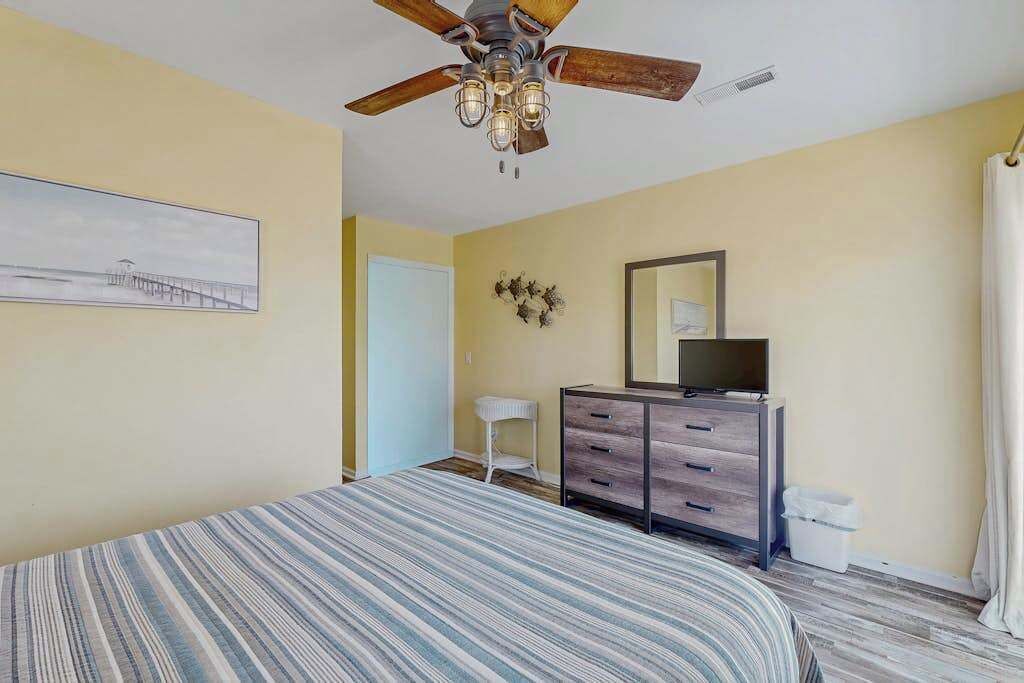 Ein Schlafzimmer mit Bett, Deckenventilator, Kommode, Spiegel, gerahmtem Bild an der Wand und einer Tür.