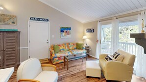 Ein Wohnzimmer mit einem farbenfrohen Sofa, einem beigen Sessel, einem Holztisch und einer weißen Tür mit der Aufschrift 'BEACH HOUSE'.