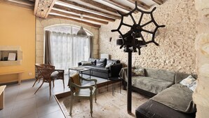 Ein gemütliches Wohnzimmer mit Steinwand, Holzbalken, einer Couch, einem Sessel, einem Tisch und einem hängenden Schiffswellenrad.