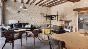 Ein rustikales Interieur mit freiliegenden Holzbalken, einer Steinwand, einem schwarzen Ledersofa, Korbsesseln, einem Holztisch und einer Küchenzeile.