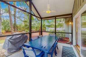 Ein überdachter Außenbereich zum Essen mit Tisch und Stühlen, einem Deckenventilator und großen Fenstern mit Blick auf Bäume und einen Zaun.