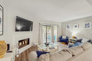 A living room with a fireplace, a sofa, a coffee table, a TV, and a balcony with outdoor furniture. - Modern Coastal Retreat | Walk to Beach | Pool & Tennis | Pet-Friendly (Hilton Head)