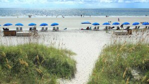 Une plage bordée de rangées de parasols bleus et de chaises longues, un ciel dégagé et l’océan en toile de fond.