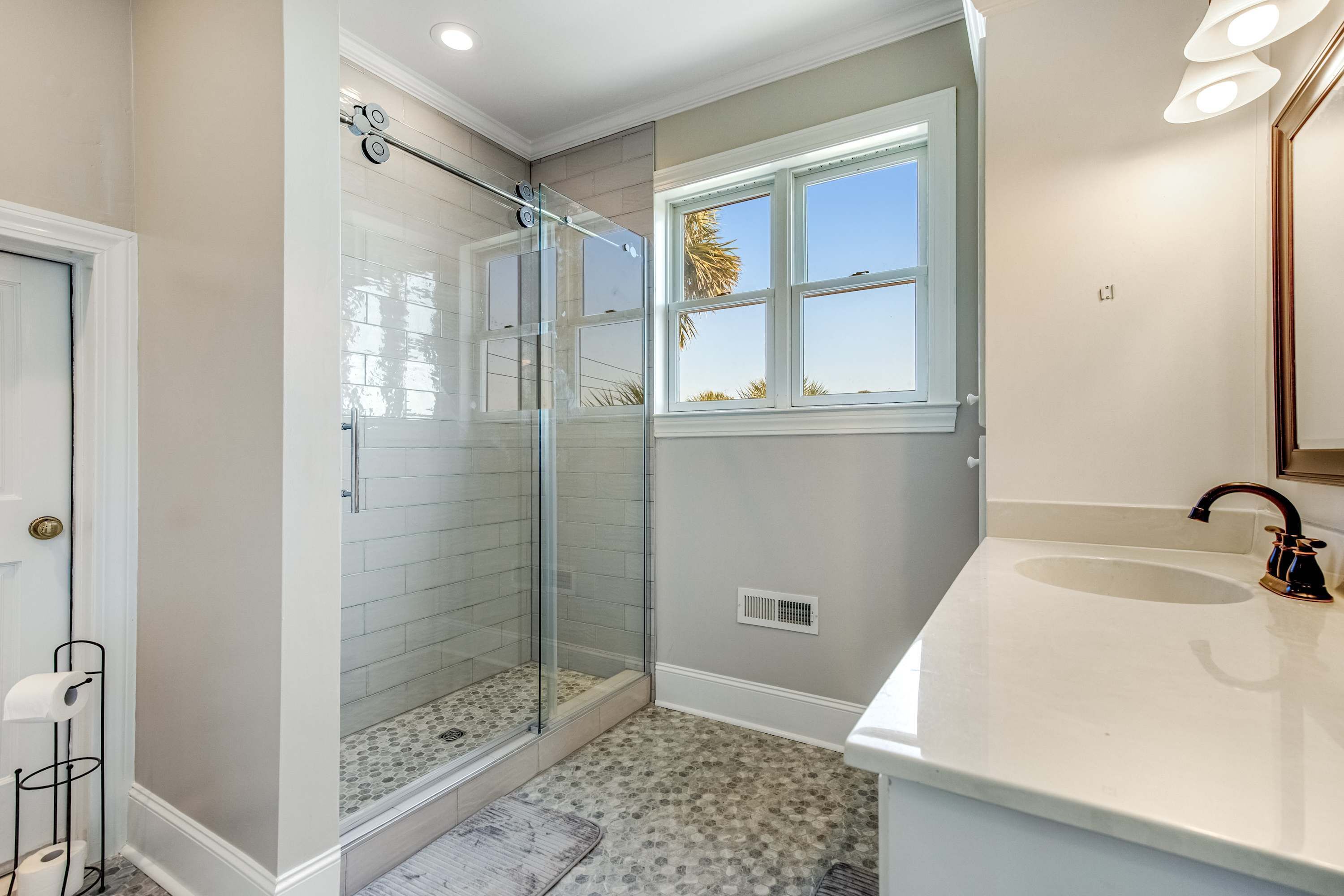 Une salle de bain avec une douche à parois de verre, un lavabo blanc et une fenêtre donnant sur des palmiers.