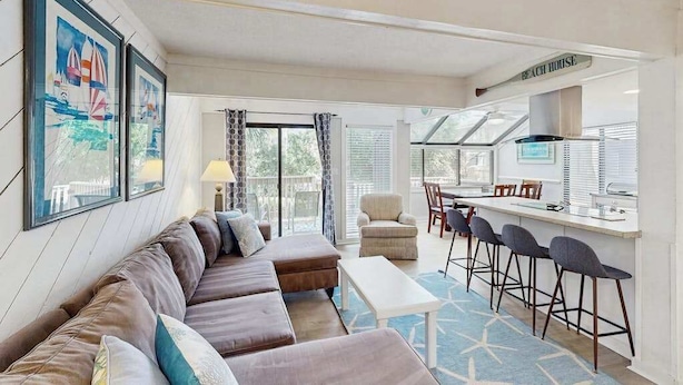 A modern living room with a sofa, a dining area with bar stools, and a kitchen area with a sign reading 'BEACH HOUSE'. - Sunny Villa with Spacious Decks, Al Fresco Dining, & Shared Pool - Nearby Beach Access (Hilton Head)