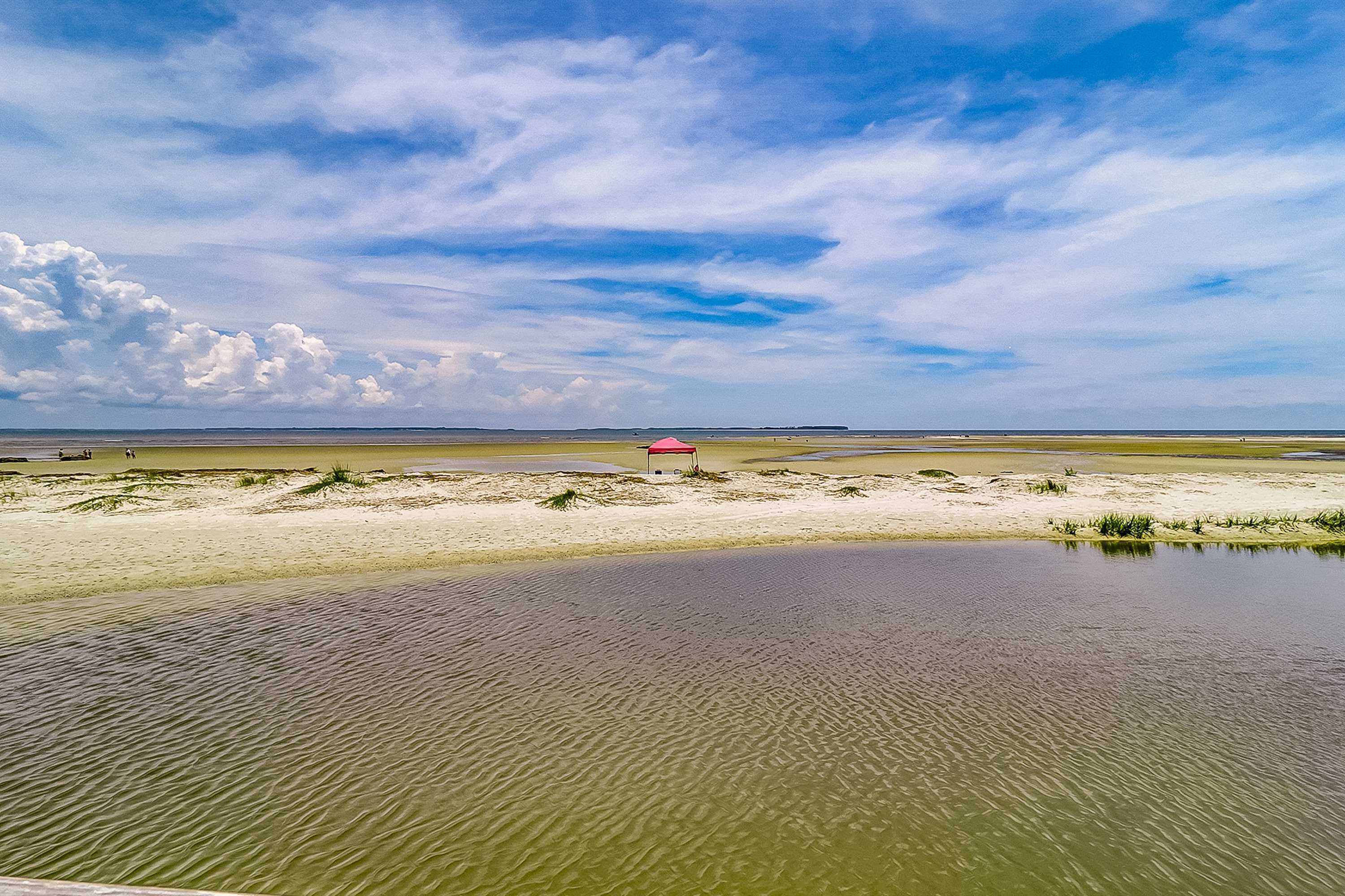 Un paisaje costero con una playa arenosa, una masa de agua pequeña y una estructura roja a lo lejos.