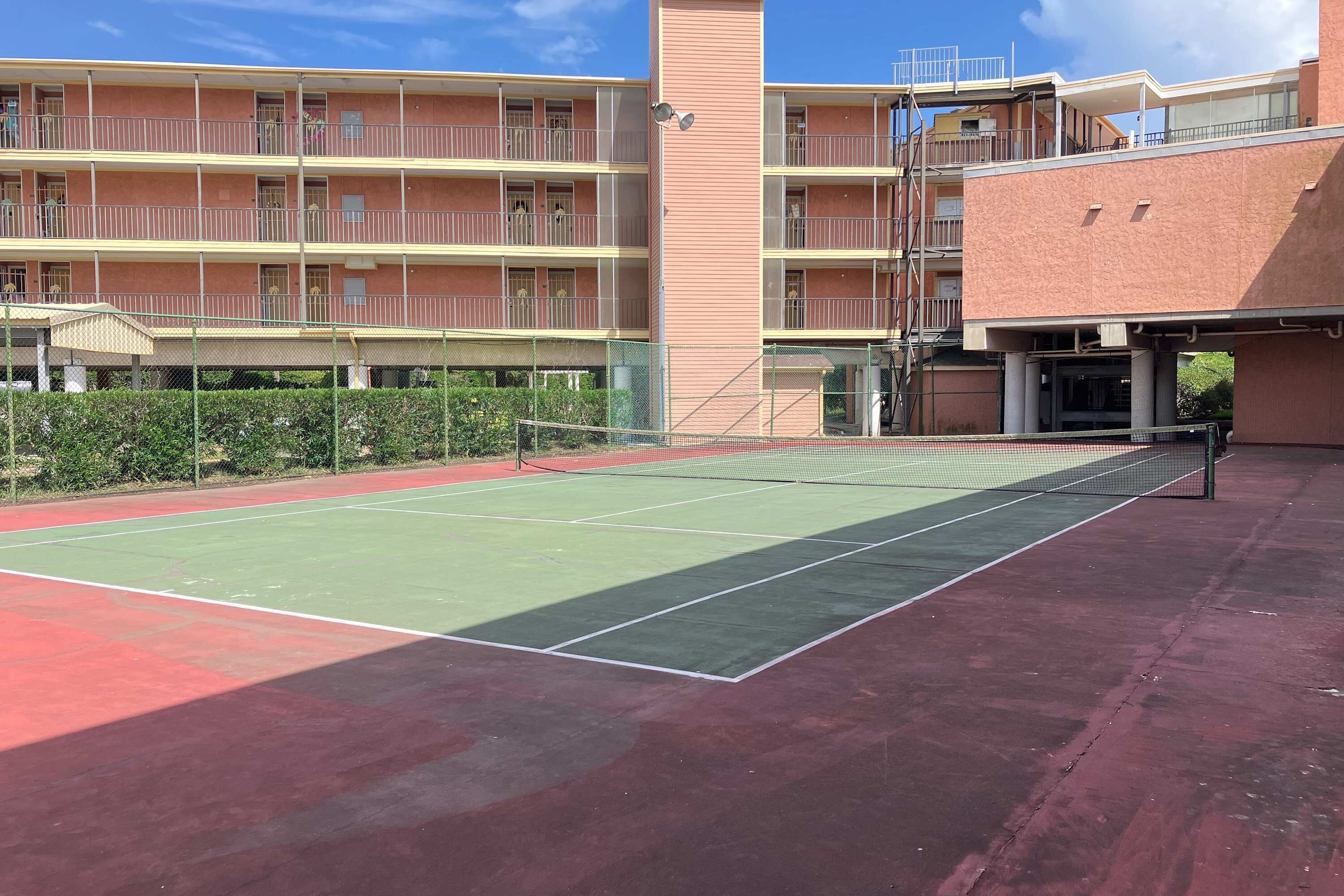 En tennisbane med net, omgivet af et hegn, og ved siden af en høj bygning med altaner.