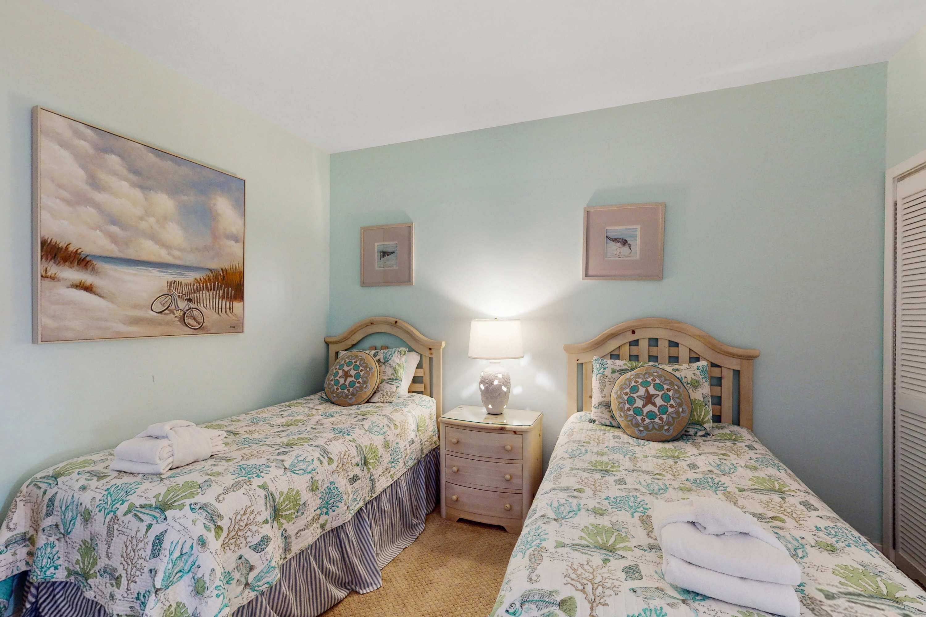 Una habitación con dos camas, una mesita de noche con lámpara, un cuadro en la pared y una ventana con persianas.