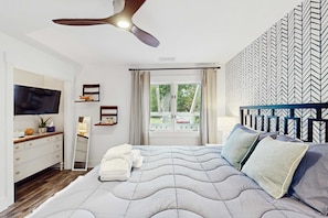 A bedroom with a bed, a ceiling fan, a TV, a dresser, a window with curtains, and a patterned wall. - Cheerful condo with shared pool, close to beach, golf, tennis and shopping (Hilton Head)