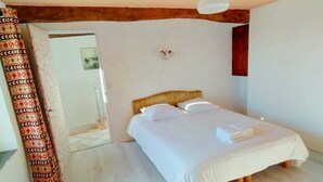 Quarto com cama, vigas de madeira, cortina estampada e porta que dá para outro cômodo.