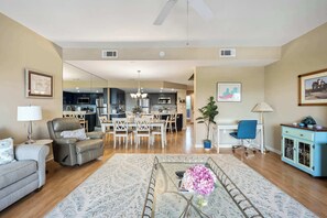 Un vaste salon comprenant un canapé, un fauteuil et une table basse, un coin repas avec des chaises et un lustre, et une cuisine en arrière-plan.