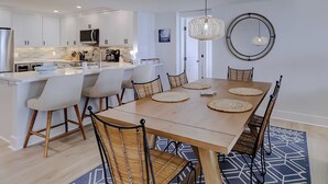 Una cocina moderna con armarios blancos, una isla central y una zona de comedor con mesa de madera y sillas de mimbre.