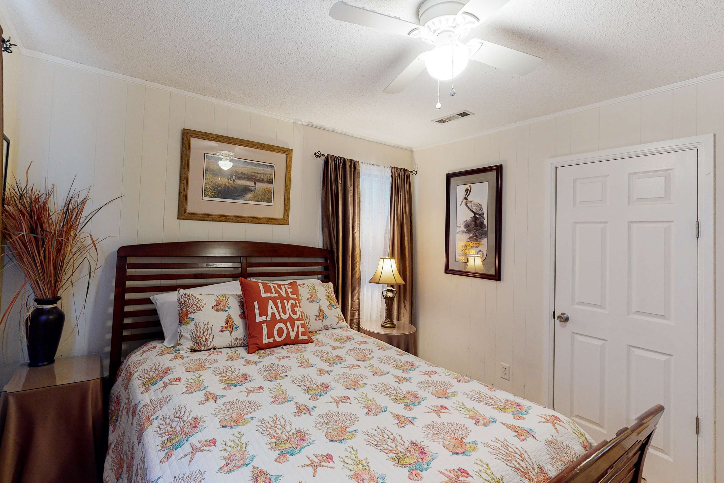 A bedroom with a bed, a ceiling fan, a nightstand, a lamp, a framed picture, a door, and a vase with dried plants.