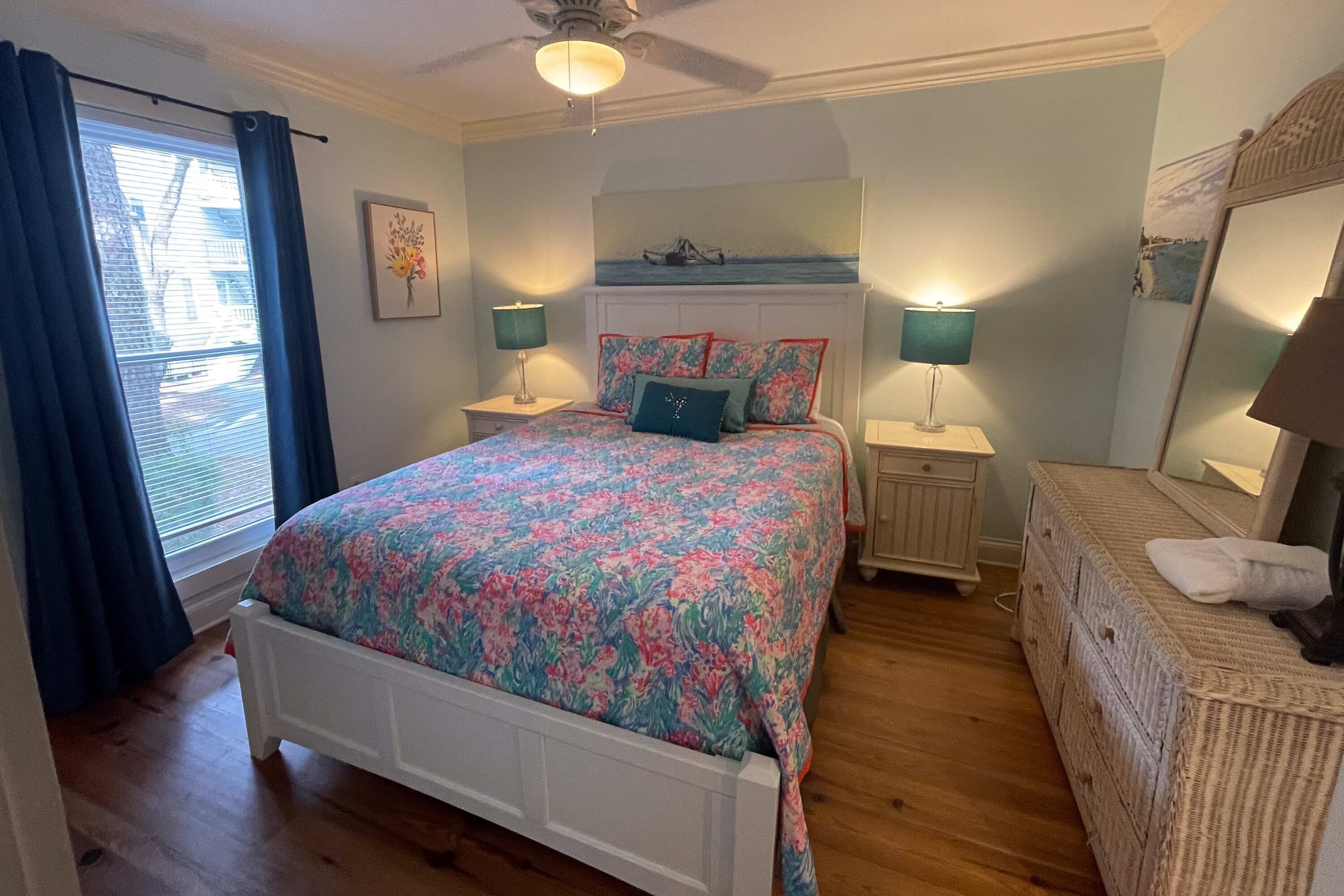 Une chambre à coucher comprenant un lit, deux tables de chevet, une commode, un ventilateur de plafond, une fenêtre avec des rideaux et un miroir fixé au mur.