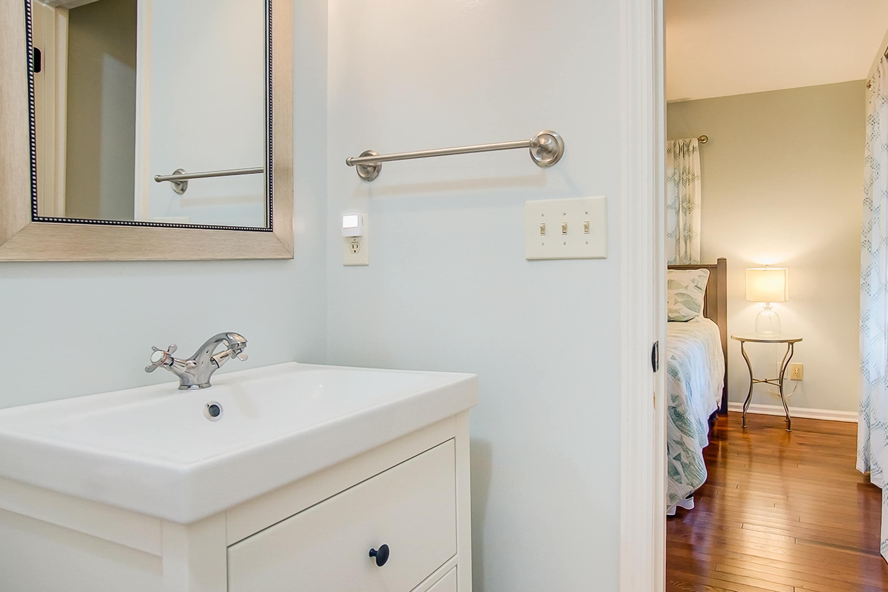 A bathroom with a white sink, a mirror, a towel rack, and a bedside table with a lamp.