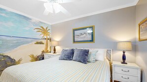 Una camera da letto con un letto, due comodini, una ventola a soffitto, un affresco raffigurante una spiaggia e un quadro appeso al muro.