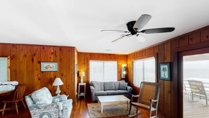 Ein Zimmer mit Holzwänden, einem Deckenventilator, einer Couch, einem Schaukelstuhl, einem Couchtisch und Blick durch große Fenster.