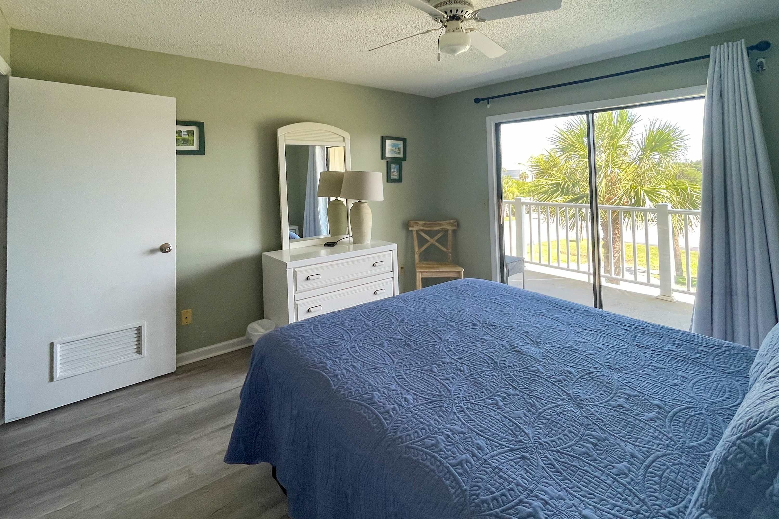 Une chambre à coucher comprenant un lit, une commode, un miroir, un ventilateur de plafond et un balcon avec vue sur les palmiers.