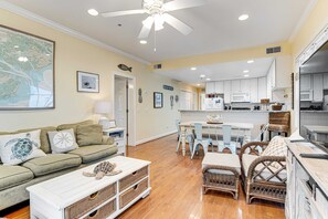 A living room with a sofa, coffee table, and wicker chairs. There is a dining area with a table and chairs. The kitchen is visible with white cabinets and a refrigerator. - Top-floor 3rd-row condo with balcony, AC, outdoor pool & beach access (Edisto Beach)