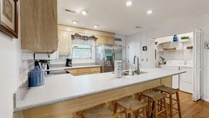 A modern kitchen with a central island, bar stools, a refrigerator, and a washing machine. - Steps to the Beach & Shops Home on a Tidal Creek w/ Two Decks, AC & WiFi (Edisto Beach)