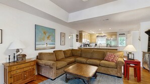 A living room with a sectional sofa, a wooden coffee table, a wooden cabinet, and a kitchen in the background. - Steps to the Beach & Shops Home on a Tidal Creek w/ Two Decks, AC & WiFi (Edisto Beach)