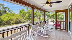 A covered porch with rocking chairs, a ceiling fan, and large windows offering a view of trees. - Steps to the Beach & Shops Home on a Tidal Creek w/ Two Decks, AC & WiFi (Edisto Beach)