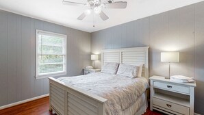 Ein Schlafzimmer mit einem Bett, Nachttischen, einem Deckenventilator, einem Fenster mit Jalousien und einem Beistelltisch mit Lampe.