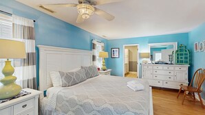 Un dormitorio con cama, mesitas de noche, cómoda, silla, ventilador de techo y puerta que da a otra habitación.