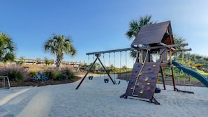 A playground with a swing set and climbing wall, surrounded by sand and palm trees. - Oceanfront villa with WiFi, full kitchen, pool, & hot tub - beach nearby (Hilton Head)