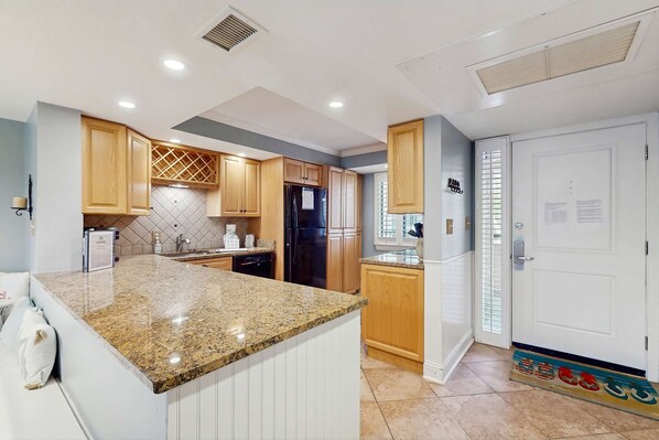 A kitchen with granite countertops, wooden cabinets, a refrigerator, and a white door. - Oceanfront villa with WiFi, full kitchen, pool, & hot tub - beach nearby (Hilton Head)