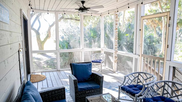 Un portico coperto con mobili in vimini, una ventola a soffitto e ampie finestre che si affacciano su un giardino.