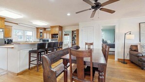 Una cocina amplia con armarios de madera, un comedor con una mesa grande y sillas de madera, un ventilador de techo y vistas al salón.
