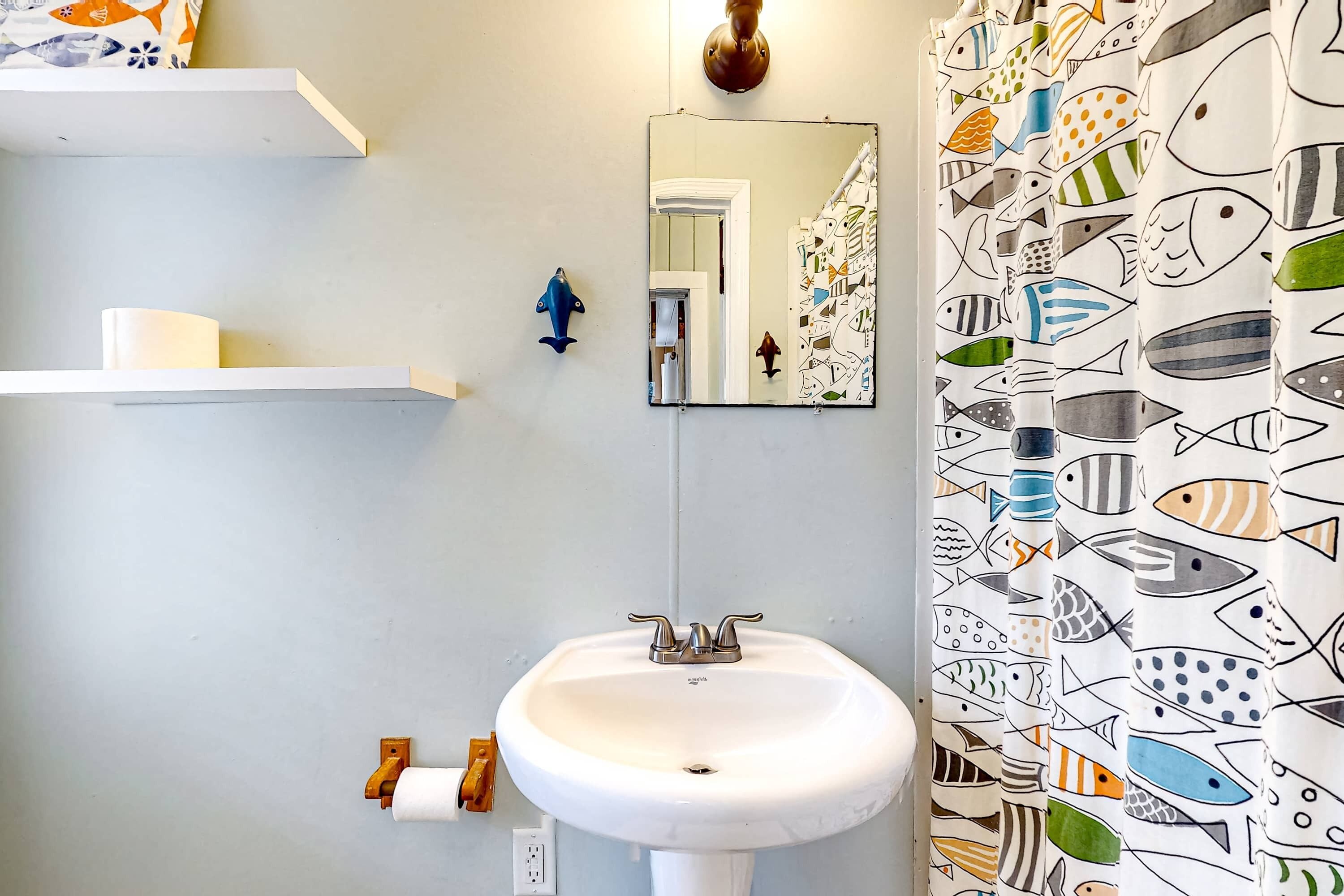 Un baño con lavamanos blanco, espejo, luz en forma de pez azul y cortina de ducha con estampado de peces.