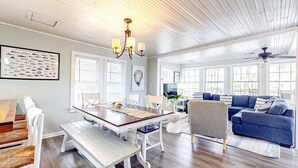 A dining area with a wooden table and white chairs, a living room with a blue sofa and striped armchair, and a ceiling fan. - Beautiful home across from the sea with balcony & open layout - dog-friendly (Edisto Beach)