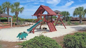 A playground with a slide and climbing structures, surrounded by sand and palm trees. - Family-Friendly Townhome with Lagoon View, Shared Pool, Hot Tub & Tennis (Hilton Head)