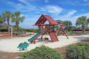 A playground with a slide and climbing structures, surrounded by sand and palm trees. - Family-Friendly Townhome with Lagoon View, Shared Pool, Hot Tub & Tennis (Hilton Head)