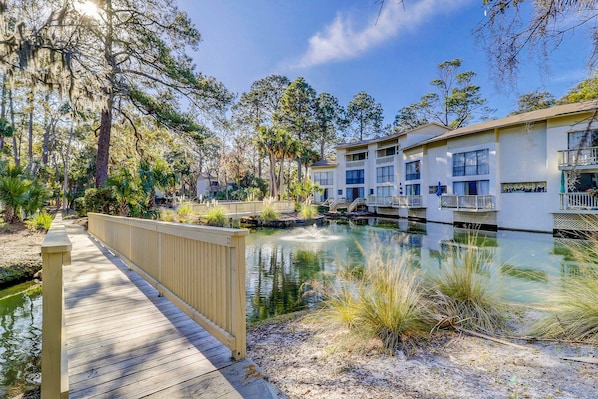 A wooden bridge over a pond with buildings in the background. - Beach access condo with deck, pool, washer dryer, and AC - snowbird-friendly (Hilton Head)