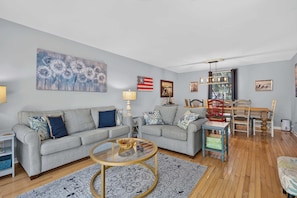A living room with two grey sofas, a coffee table, wooden flooring, and a dining area with chairs and a table. - Beach access condo with deck, pool, washer dryer, and AC - snowbird-friendly (Hilton Head)
