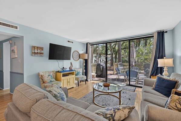 A living room with a sofa, armchairs, a coffee table, a TV, and a view of an outdoor patio with chairs and umbrellas. - Beach access condo with deck, pool, washer dryer, and AC - snowbird-friendly (Hilton Head)