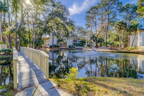 A wooden bridge over a calm water body, surrounded by trees and buildings with a clear sky. - Beach access condo with deck, pool, washer dryer, and AC - snowbird-friendly (Hilton Head)