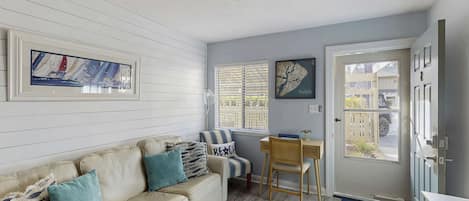 Una sala de estar acogedora con un sofá color beige, una silla a rayas, un escritorio de madera y una puerta que da al exterior.