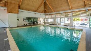Una piscina interior con techo de madera, paredes de vidrio transparente y suelo de baldosas.