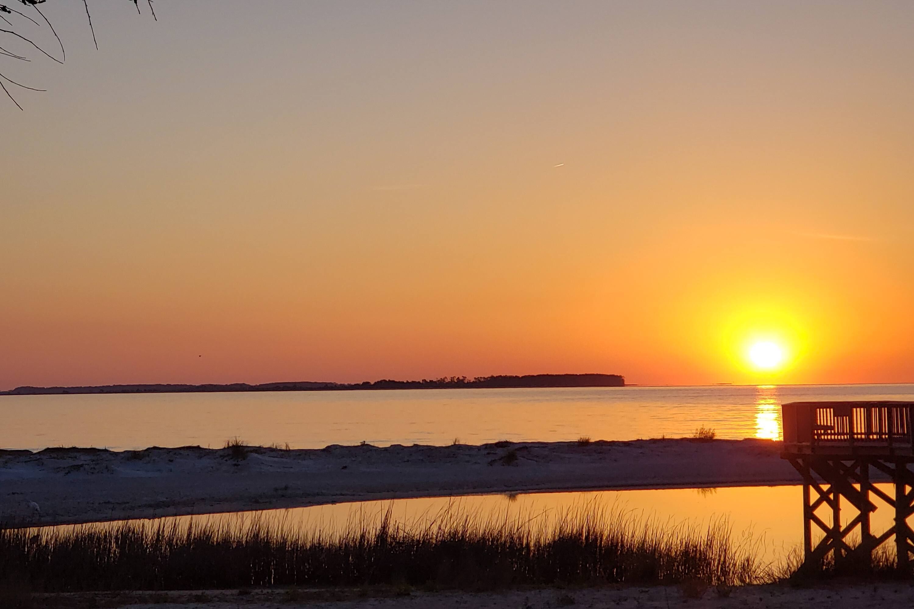 A sunset over a calm body of water with a wooden pier and sand dunes in the foreground.