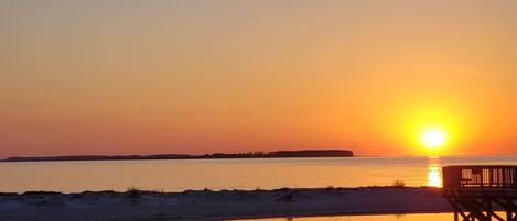 A sunset over a calm body of water with a wooden pier and sand dunes in the foreground.
