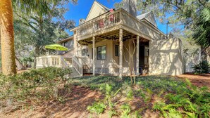 A two-story house with a balcony, a covered porch, and a wooden fence. - Coastal Villa with Spacious Deck, Pool & Walk to Beach (Hilton Head)