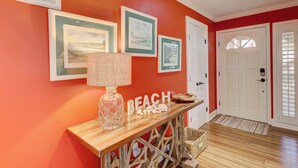 A hallway with a wooden console table, framed pictures, a 'BEACH' sign, and a white door. - Coastal Villa with Spacious Deck, Pool & Walk to Beach (Hilton Head)