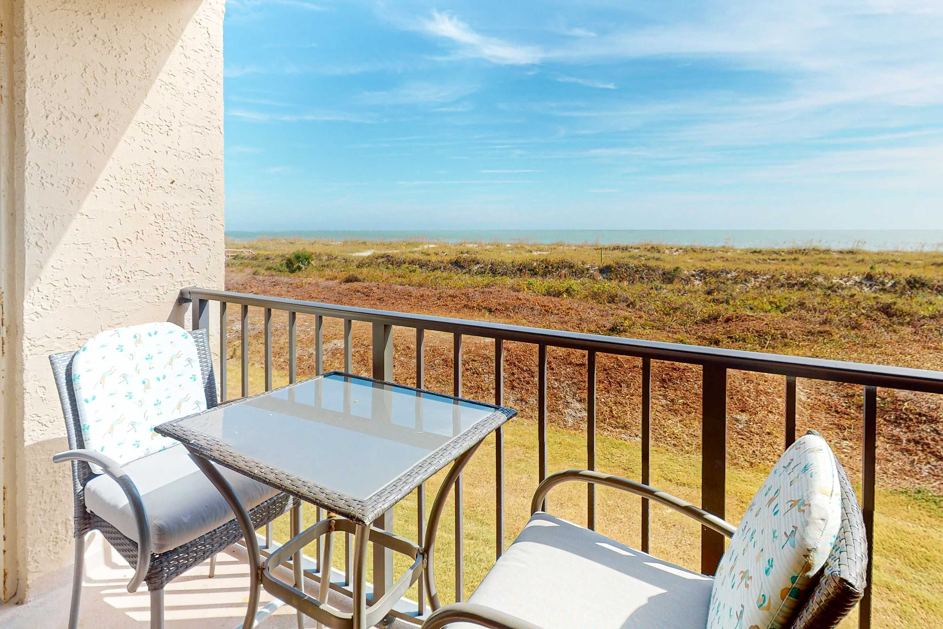 A balcony with a table and two chairs, overlooking a coastal landscape.